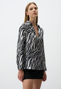 Blusa a stampa zebra bianca e nera in tessuto liscio e lucido. Caratterizzata da un collo a V e maniche lunghe, con una vestibilità ampia.