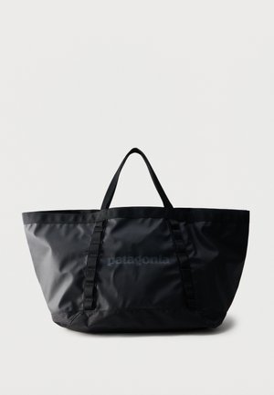 Čierna taška Patagonia typu tote s krátkymi rukoväťami a vertikálnymi slučkami na prednej strane, na jednoliatom svetlosivom pozadí.