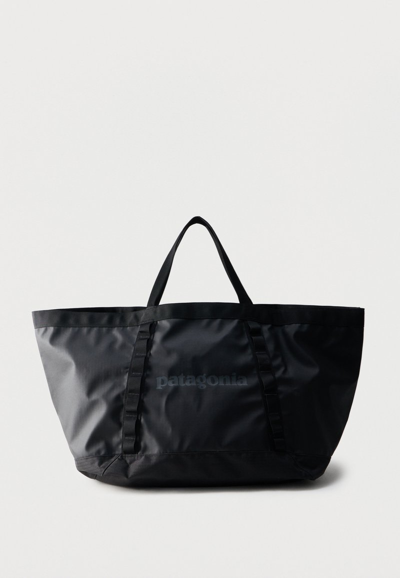 Μαύρη τσάντα tote Patagonia με κοντά χερούλια και κάθετες θηλιές μπροστά, σε απλό ανοιχτό γκρι φόντο.