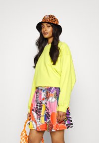 Neon zielony, oversize sweter, kolorowa mini spódnica z geometrycznymi i zwierzęcymi wzorami, pomarańczowy kapelusz w panterkę, pomarańczowa torba.