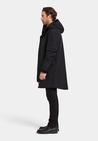 Svart vattentät rock med huva, rak skärning, sidofickor och långa ärmar. Bärs över svarta byxor och stövlar.