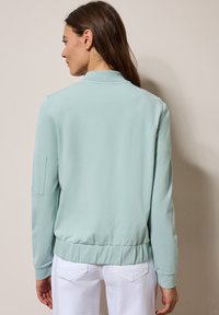 Mintgrüne Bomberjacke mit gerippten Bündchen und Saum, die über einer glatten Textur und Seitentasche verfügt, getragen über weißen Hosen.