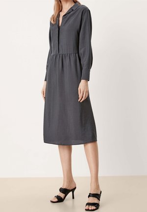 Vrouw in een midi-lange, donkergrijze jurk met lange mouwen en een kraag, en zwarte sandalen met hak, tegen een effen achtergrond.