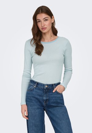 Jeune femme aux cheveux bruns ondulés portant des boucles d'oreilles anneau, un haut texturé à manches longues bleu clair et un jean bleu taille haute, une main dans la poche.