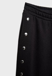Pantalon en tissu noir avec une taille élastique comportant une rangée verticale de six boutons-pression argentés brillants le long de la couture latérale.