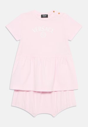 Růžové bavlněné šaty s rozšířeným spodním dílem, zdobené logem "VERSACE" a dekorativním límcem se dvěma zlatými patentkami. Pod nimi jsou připojené šortky.