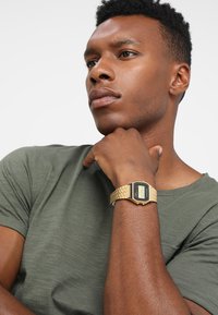 Montre en or numérique avec un cadran rectangulaire, bracelet en maillons métalliques et écran noir. La personne porte une chemise verte, la main reposant sous son menton.