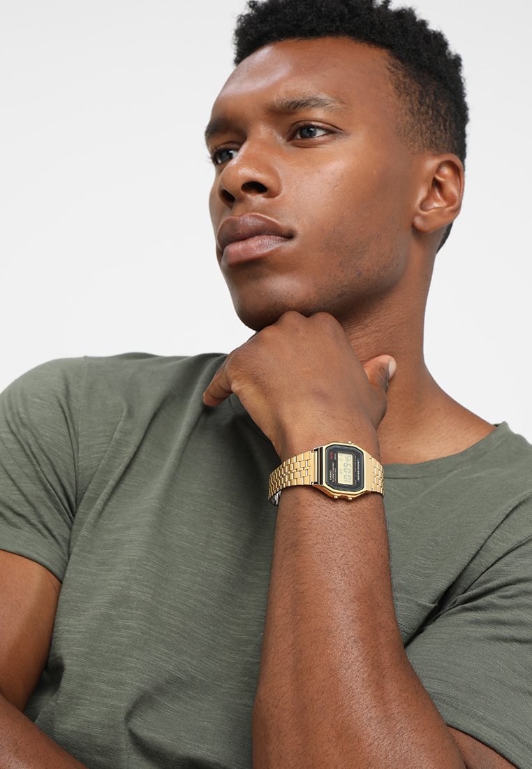 Montre en or numérique avec un cadran rectangulaire, bracelet en maillons métalliques et écran noir. La personne porte une chemise verte, la main reposant sous son menton.