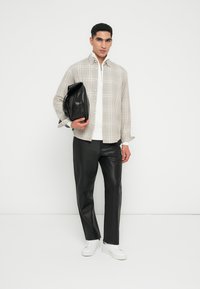 Camisa de cuadros clara con botones, camiseta blanca debajo, pantalones de cuero negros y zapatillas blancas. El modelo sostiene un bolso de cuero negro.