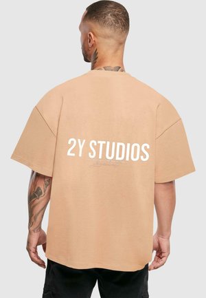 Overdimensjonert beige bomull t-skjorte med "2Y STUDIOS" i hvitt på baksiden. Har rund hals og korte ermer. Glatt tekstur.
