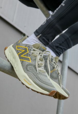 Osoba nosząca szare i żółte buty do biegania New Balance, białe skarpetki oraz czarne spodnie zapinane na zamek, opiera stopy na metalowym pręcie.