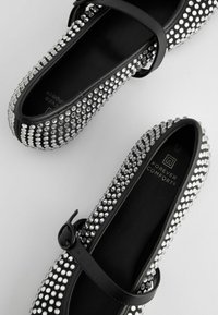 Des chaussures plates noires ornées de strass argentés, avec une bride brillante. L'intérieur présente un revêtement noir et un logo. Texture lisse.