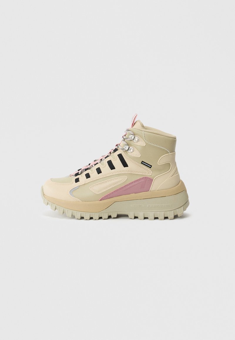 Bota de senderismo impermeable en beige y rosa con suela de grosores gruesos, lazos de cordones negros y soporte para el tobillo, mostrada en perfil lateral sobre fondo blanco.