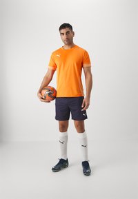Oranž T-särk lühikeste varrukatega valge logoga, tumesinised lühikesed püksid, valged sokid ja mustad spordijalatsid, hoides mustrilist jalgpalli.