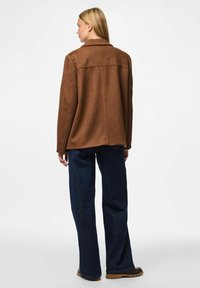 Smeđi veličanstveni jakna od suedea s opuštenim krojem, koja ima ovratnik i vidljive šavove, u kombinaciji s tamnoplavim širokim trapericama i crnim čizmama.