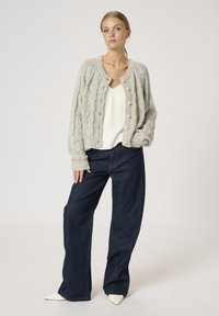Hellgrauer Strickcardigan mit strukturiertem Kabelmuster, mit Knopfverschluss, getragen über einem weißen Oberteil und dunkelblauen weit geschnittenen Jeans.