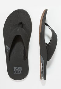 Zwarte flip-flops met een gestructureerde rubberen zool, elastische banden en een contrasterende bruine voetbed. Heeft een transparante accent aan de zijkant.