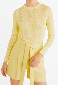 Top a maniche lunghe di colore giallo chiaro lavorato a maglia con un motivo a zig zag, abbinato a pantaloni corti in vita alta con cintura annodata e tasche.