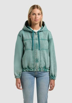 Tânără purtând o jachetă glugă cu fermoar de culoare teal, cu șnururi și buzunare frontale, asortată cu blugi albastru deschis, stând într-un cadru simplu.