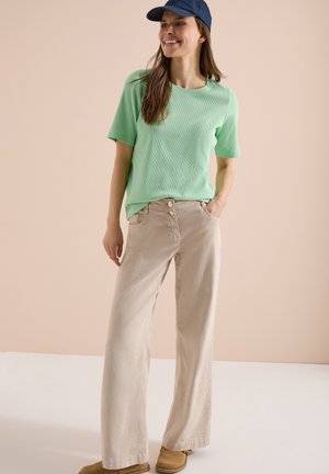 Femme portant une chemise à manches courtes vert menthe, un pantalon large beige, des chaussures couleur cuir clair et une casquette bleu marine, debout avec une main dans la poche.