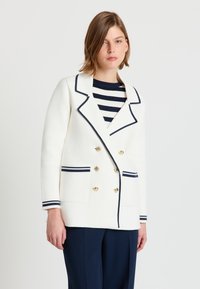 Frau trägt ein weißes, zweireihiges Blazer mit marineblauen Besätzen und goldenen Knöpfen über einem marineblau-weiß gestreiften Hemd, kombiniert mit marineblauen Hosen.