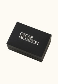 Musta suorakulmainen laatikko, jossa on sileä pinta, ja siinä on hopeinen "OSCAR JACOBSON" -logo. Laatikko on suunniteltu säilytystä tai pakkauksia varten.