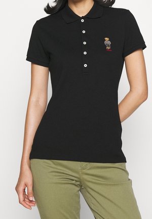 Kvinde iført en sort kortærmet polo-shirt med en lille broderet bjørn på brystet og olivengrønne bukser.