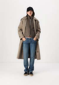 Trench beige sobre una chaqueta acolchada marrón, jeans azules y zapatillas negras. La gorra es negra. Conjunto en capas casual.