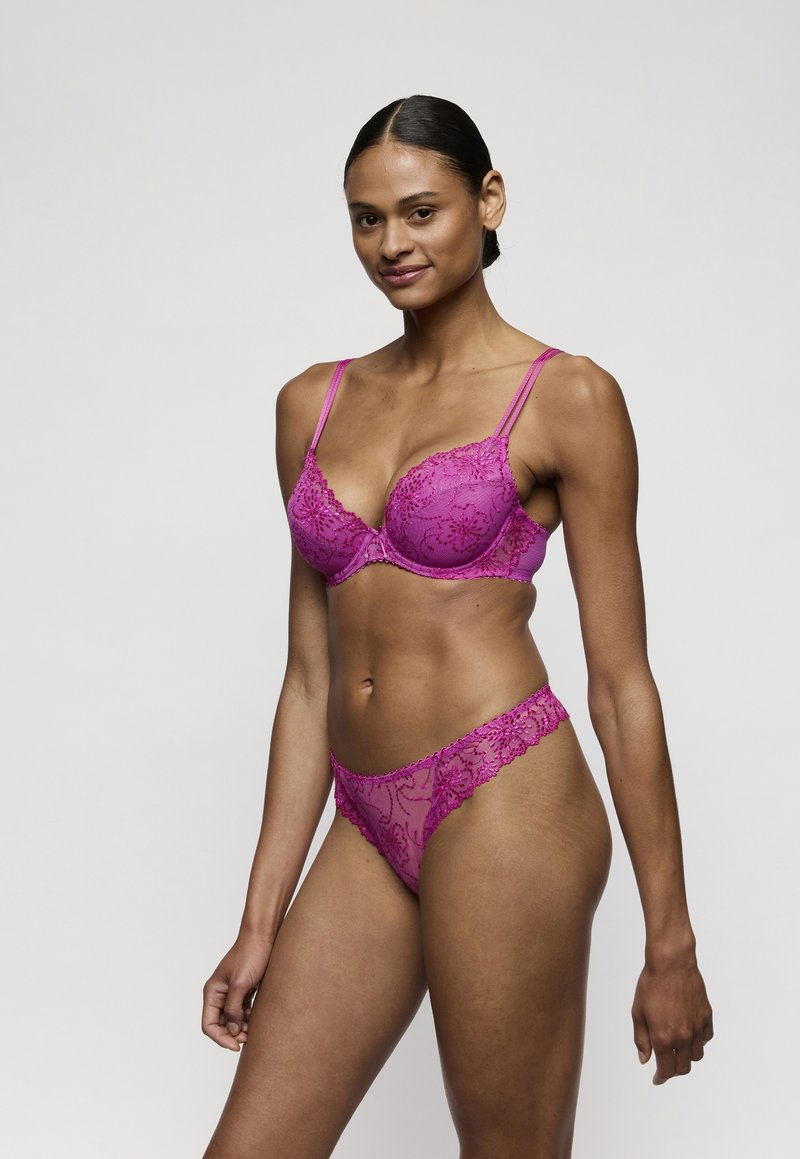 Frau mit dunklen Haaren zu einem Dutt gebunden, trägt einen passenden lila Spitzen-BH und Spitzen-Slips, steht vor einem schlichten hellen Hintergrund und lächelt leicht.