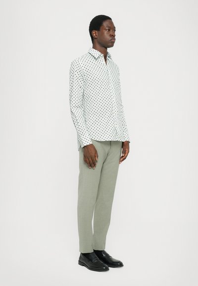 Long-sleeve shirt in white with black polka dots, paired with light green trousers. Model wears black shoes, standing against a plain background.
