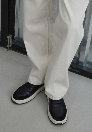 Zwarte leren sneakers met een gestructureerde afwerking, witte zool en veters. Gepaard met losse, lichtbeige broek.