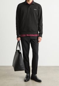 Černá mikina s kapsou, logo na hrudi, vrstvená přes vínovou košili. Černé kalhoty, lesklé boty a velká černá taška na rameno.