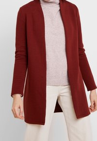 Cardigan côtelé couleur marron avec ouverture sur le devant, manches longues et tissu texturé. Porté sur un col roulé rose clair et associé à un pantalon de couleur claire.