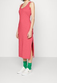 Rosa, ärmlös midiklänning med sidoklipp, tillverkad av ett texturerat tyg. Kombinerad med vita sneakers och gröna, lager-på-lager-strumpor för kontrast.