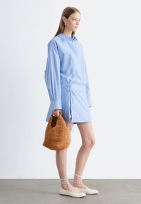 Jeune femme portant une mini-robe à manches longues bleu clair, tenant un sac à main tressé marron et portant des ballerines blanches à lacets.