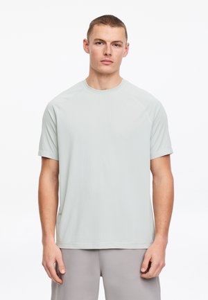 Joven con cabello corto que lleva una camiseta gris claro de manga corta con cuello redondo y pantalones grises, de pie contra un fondo blanco liso.