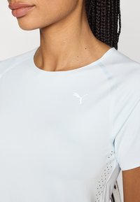 Ljust blå sporttop med kort ärm, med en liten vit logotyp på bröstet och ventilerade sidodetaljer för andningsförmåga.