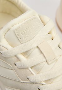 Sneaker w kolorze kremowym, wykonany z zamszu i tkaniny. Posiada płaskie sznurowadła, teksturowaną metkę oraz element haftowany po boku.