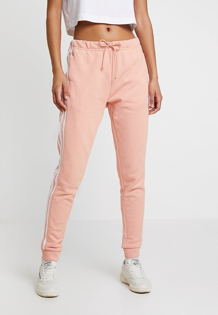Ljusrosa sweatpants med dragsko i midjan, avsmalnande ben och vita sidolinjer. Mjuk tyg med en slät yta. Skorna är vita sneakers.