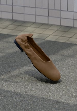 Brown soft leather flat shoe with rounded toe on gray pavement near a tiled wall.