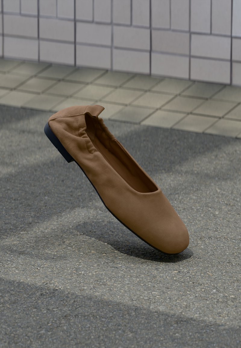 Chaussure plate en cuir souple marron avec bout arrondi sur un trottoir gris près d'un mur carrelé.