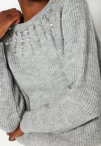 Grau gestrickter Pullover mit gerippter Textur und verziertem Ausschnitt, der mit klaren, juwelenähnlichen Akzenten in einem absteigenden Muster geschmückt ist.