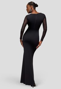 Zwarte jurk met volle lengte en lange doorschijnende mouwen, nauwsluitende silhouet, ronde halslijn en een ritsdetail aan de achterkant. Gladde textuur.