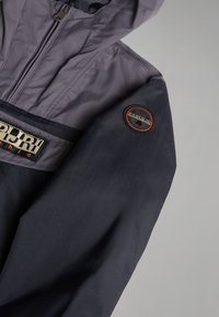 Veste gris foncé et noire avec une fermeture éclair, une capuche, un patch logo Napapijri sur la manche et une étiquette de marque en tissu rectangulaire sur la poitrine.