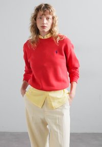 Felpa rossa con polsini a coste e un piccolo logo, indossata sopra una camicia gialla con colletto. Abbinata a pantaloni larghi beige.