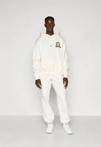 White hooded sweatshirt and matching pants made of soft fabric. Features a logo patch on the chest. Worn with white sneakers.