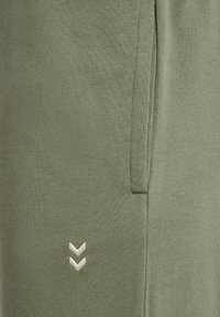 Olivegrüne Jogginghose aus weichem Stoff, mit einer Seitentasche und einem weißen Chevron-Logo, das vorne links aufgestickt ist.