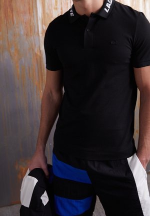 Homme portant un polo noir avec le texte "LACOSTE" sur le col, un short noir et bleu, tenant une veste noire et blanche.