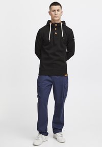 Felpa nera con cappuccio e bottoni, con lacci beige. Abbinata a pantaloni cargo blu e sneakers bianche. Design semplice.