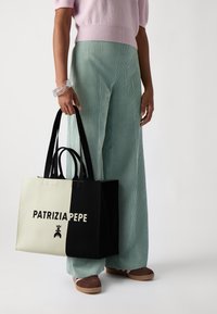 Czarna i biała torba płócienna z napisem "PATRIZIA PEPE", w dużym prostokątnym kształcie, z dwoma uchwytami i gładką fakturą. Noszona z spodniami w poziome paski w odcieniu teal.